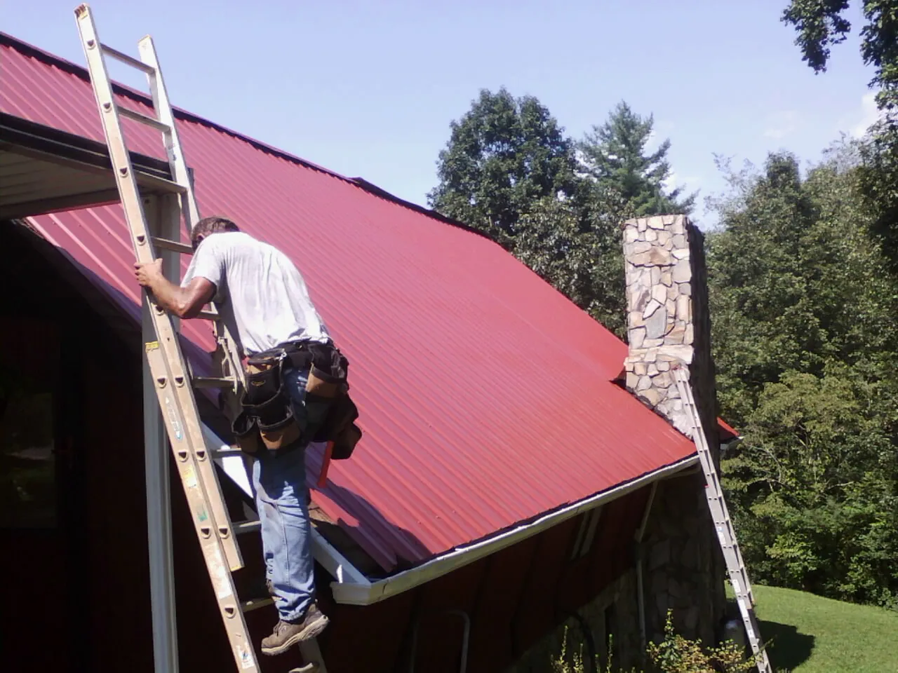 Roofer on a ladder beside a red metal roof and stone chimney — Soffit & Fascia Repair by Roofing Brothers in West Frankfort
